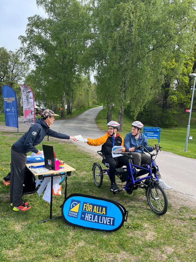 Linnéa delar ut en karta till två killar i en sorts tandemcykel. De cyklar runt banan, båda har en mindre synnedsättning.